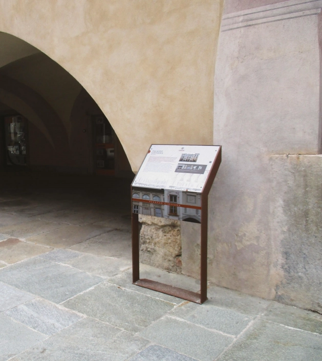 leggio-hinweistafel-07 Alt-Text: Informationsschild in einer historischen Umgebung, das Details zu einem Gebäude oder einer Sehenswürdigkeit zeigt, neben einer steinernen Wand und unter einem Bogen.