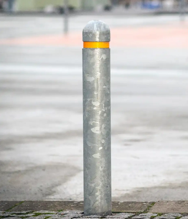 Ein grauer, zylindrischer Poller mit einer orangefarbenen Markierung oben, steht auf einem gepflasterten Boden.