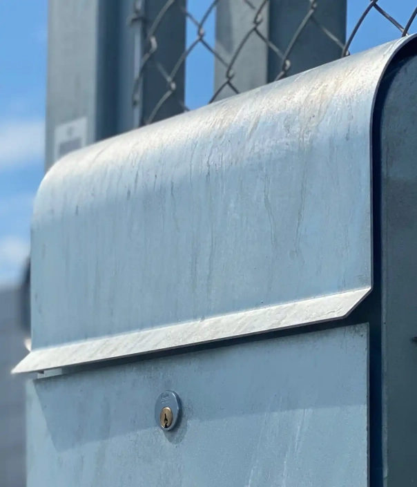 Alt-Text: Nahaufnahme eines silbernen Briefkastens mit Schloss, der an einem Zaun angebracht ist.