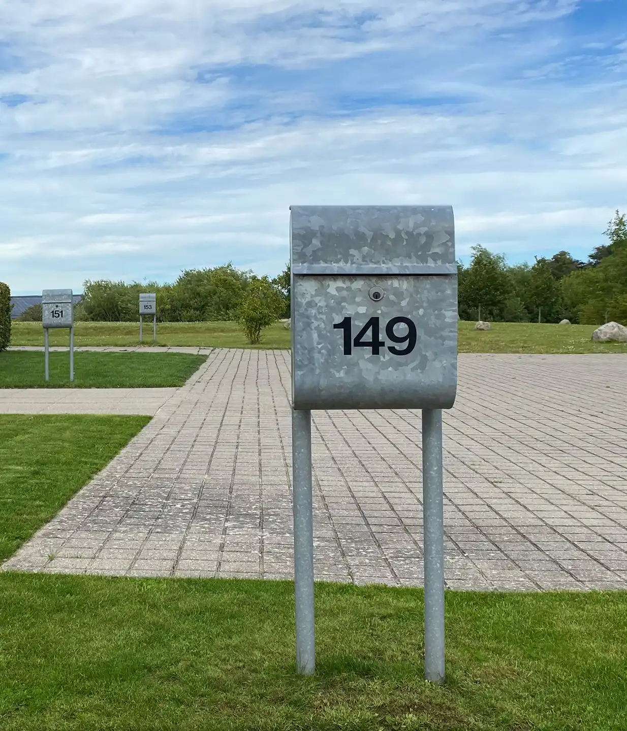 Alt-Text: Metallbriefkasten mit der Nummer 149 auf einem gepflasterten Weg, im Hintergrund weitere Briefkästen und eine grüne Landschaft.