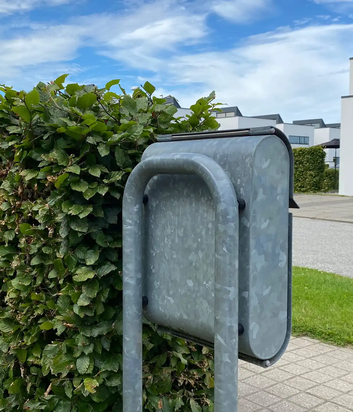 Alt-Text: "Moderner Briefkasten aus Metall, auf einem Standfuß vor einer grünen Hecke in einem Wohngebiet."