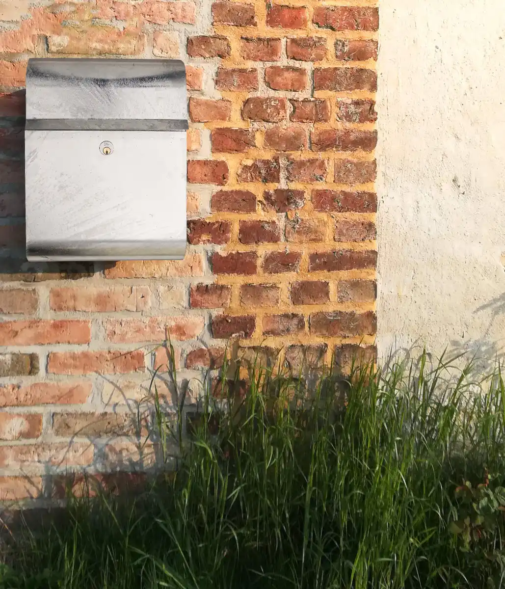 Alt-Text: Ein silberner Briefkasten hängt an einer rustikalen Ziegelmauer, umgeben von grünem Gras.