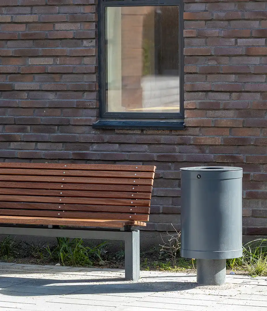 Eine Holzbank steht neben einem runden grauen Mülleimer, vor einer braunen Backsteinwand und einem Fenster.