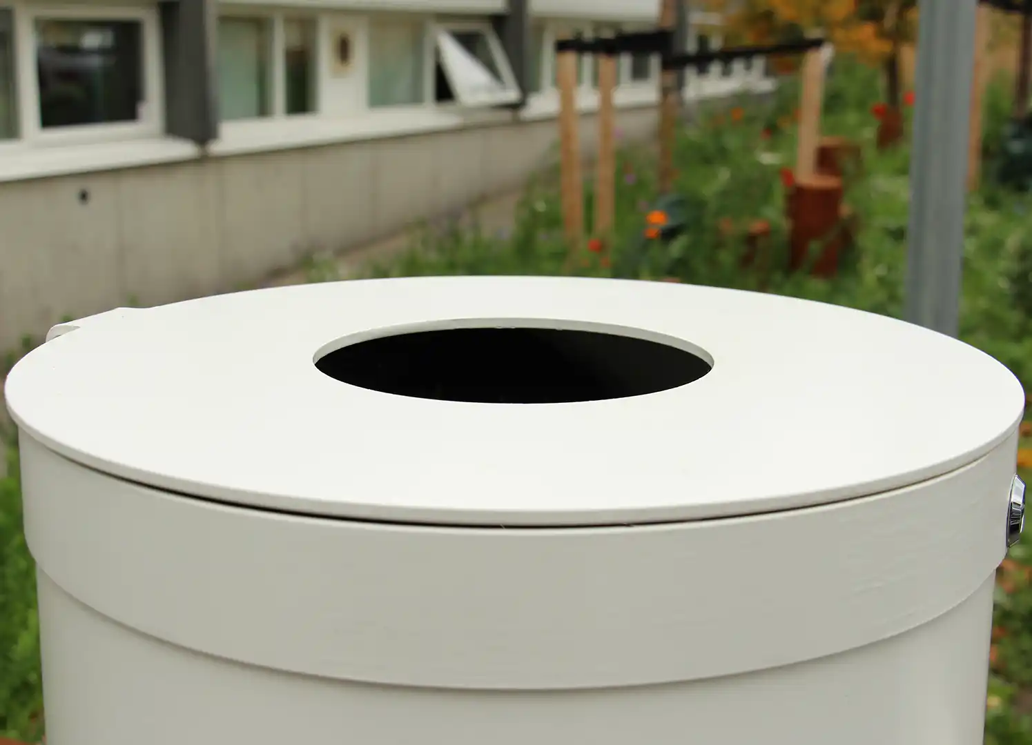 Nahaufnahme eines weißen Abfallbehälters mit runder Öffnung, im Hintergrund sind verschwommene Garten- und Gebäudestrukturen zu sehen.