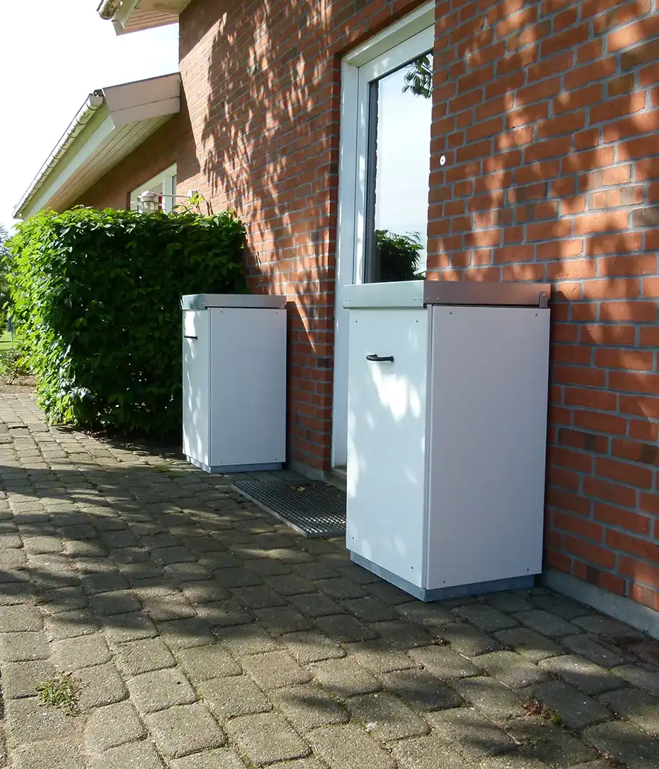 Zwei weiße Mülltonnen stehen an einer Hauswand aus roten Ziegeln, umgeben von einem Grünstreifen. Die Oberfläche der Tannen ist glatt und modern, und die Sonne wirft Schatten auf den Gehweg.