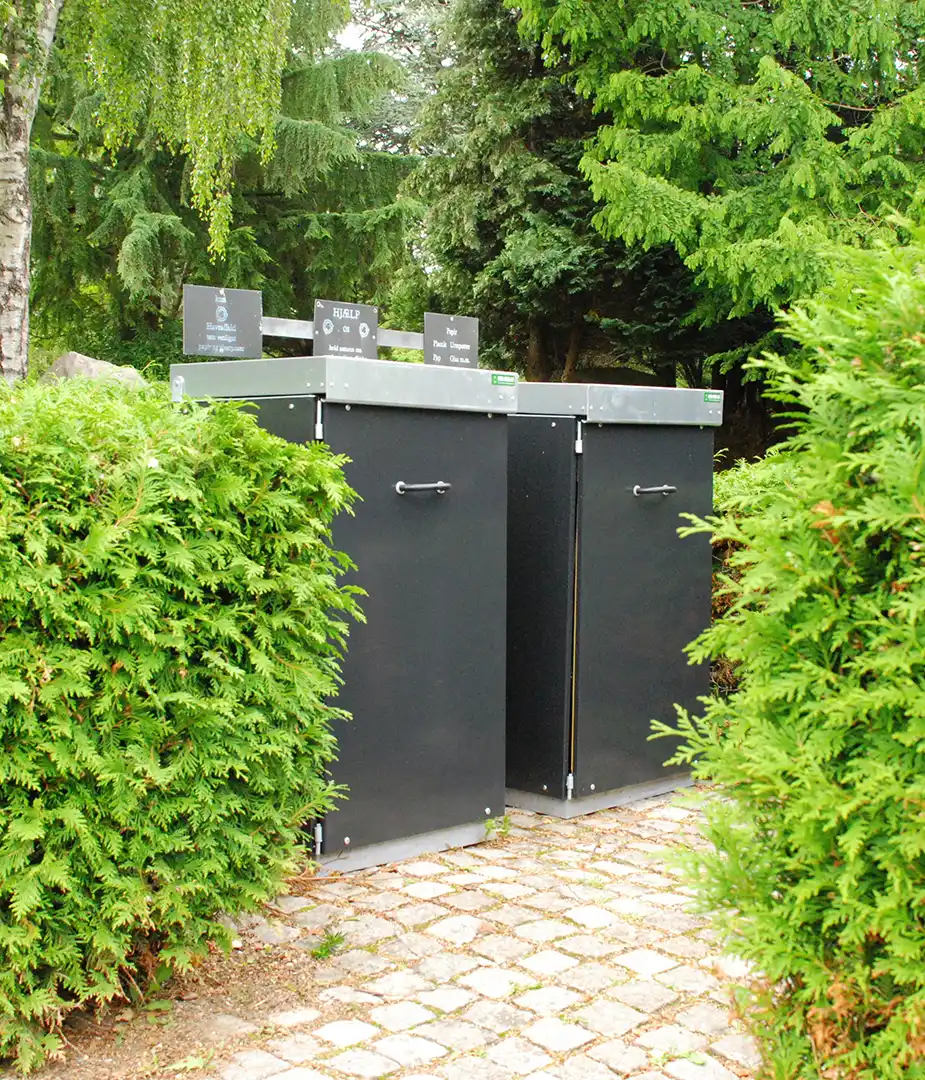 Zwei schwarz beschichtete Abfallcontainer, umgeben von dichten grünen Hecken, in einer parkähnlichen Umgebung.