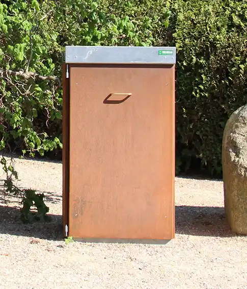 Ein rostfreier Abfalleimer aus Metall steht in einem Park auf einem Kiesweg, umgeben von Sträuchern und Steinen.
