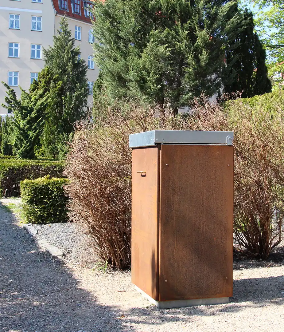 Ein rustikaler, metallener Kasten steht in einem Gartenbereich, umgeben von Sträuchern und Bäumen. Im Hintergrund sind Gebäuden sichtbar.