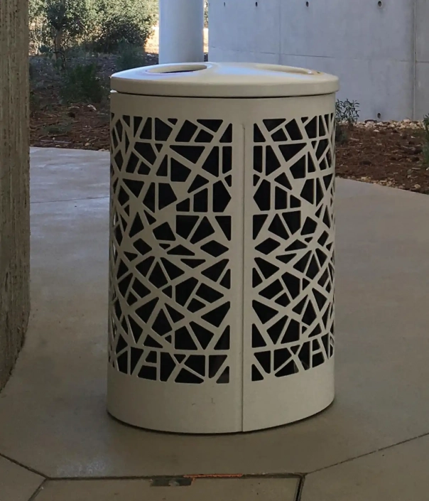 Ein moderner Abfallbehälter mit einem geometrischen Muster steht auf einem Gehweg, umgeben von grünen Pflanzen und einer minimalistischen Gebäudeoberfläche.