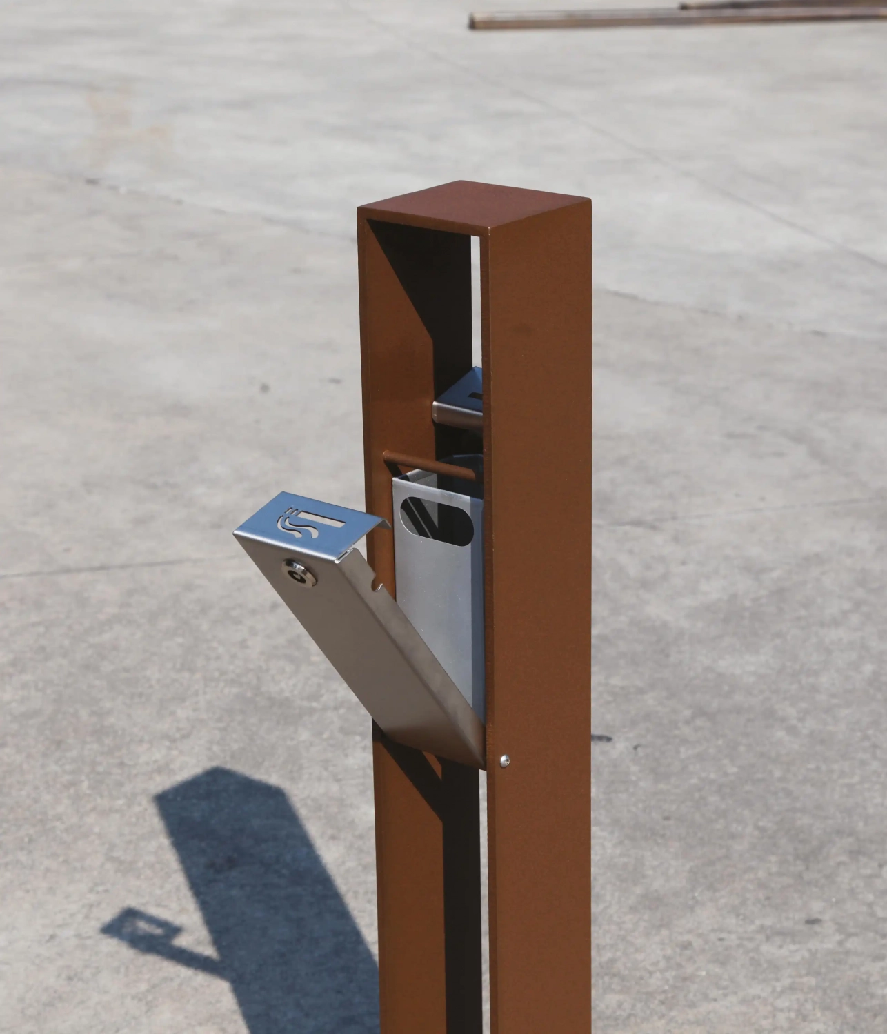 Alt-Text: "Moderner Ascher aus rostfreiem Stahl, montiert an einem robusten, braunen Metallpfosten auf einem Betonboden."