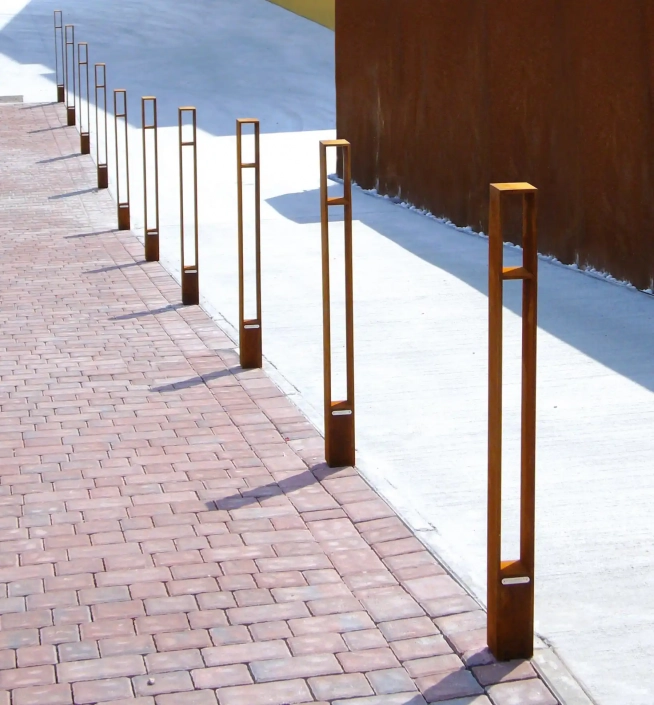 Alt-Text: Eine Reihe von schlanken, rechteckigen Pollern aus Metall, die entlang eines gepflasterten Gehwegs aufgestellt sind, mit langen Schatten, die durch das Sonnenlicht entstehen.