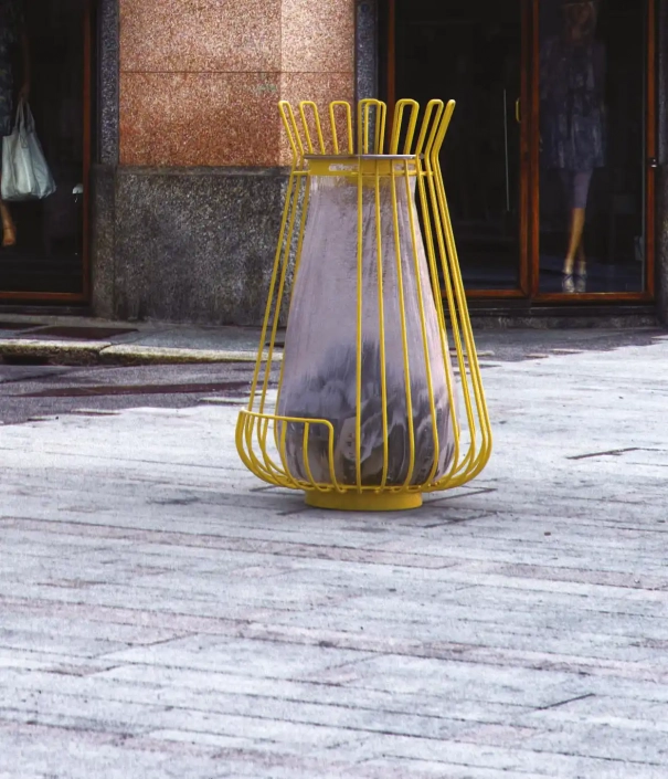 Ein ungewöhnlicher gelber Abfallkorb in Form eines Kegels steht auf einem Gehweg, umgeben von blassem Pflaster und eleganten Schaufenstern im Hintergrund.