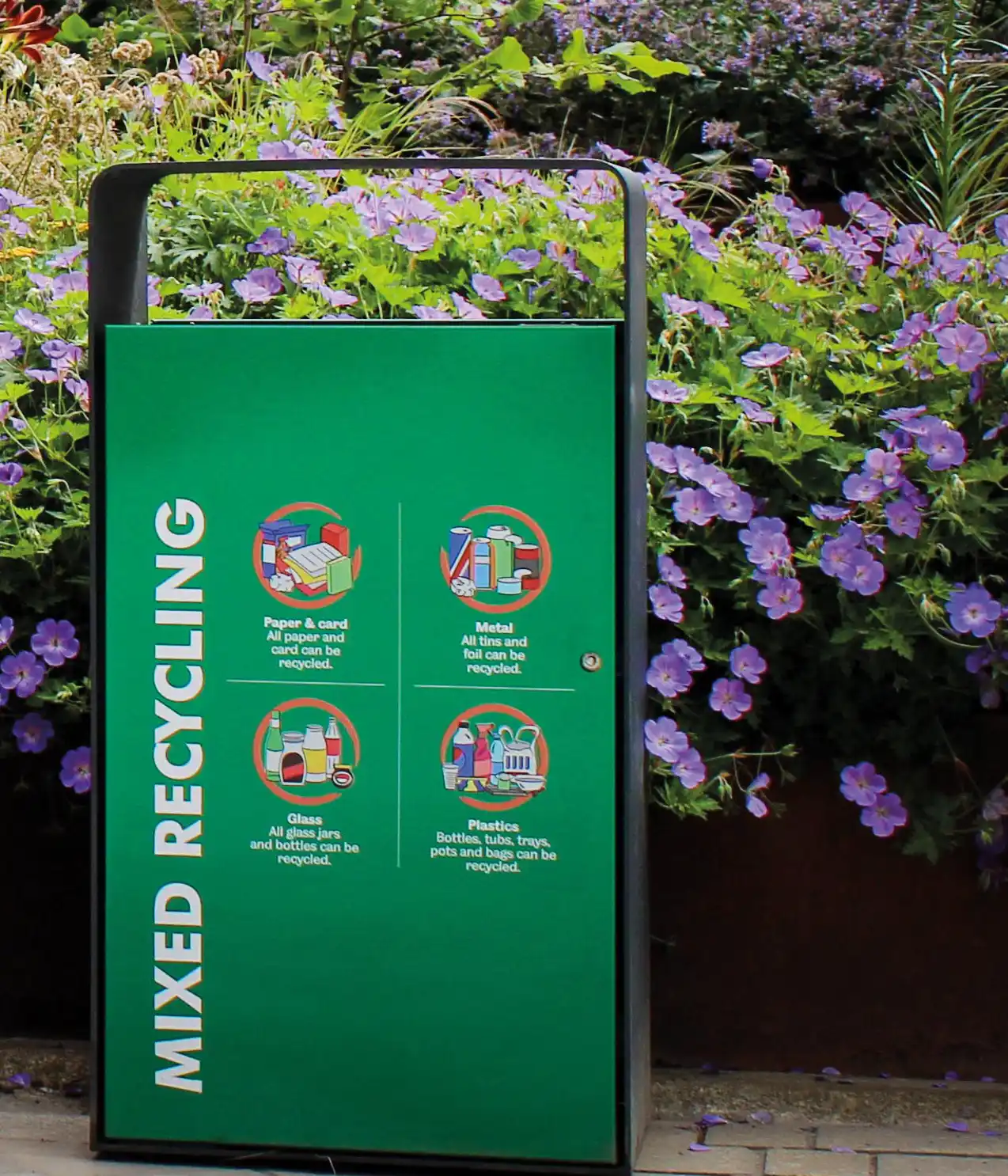 Eine grüne Recycling-Station mit dem Aufdruck "Mixed Recycling". Darauf sind Symbole und Beschreibungen für die zu recycelenden Materialien: Papier, Metall, Glas und Plastiken, umgeben von blühenden Pflanzen.