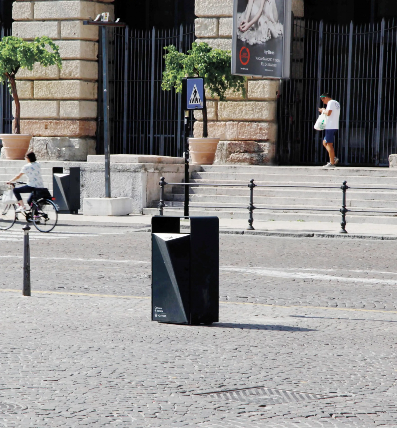Eine Straße mit einem Abfallbehälter im Vordergrund. Im Hintergrund fährt eine Person auf einem Fahrrad vorbei, während weitere städtische Elemente, wie Laternen und Pflanzkübel, sichtbar sind.