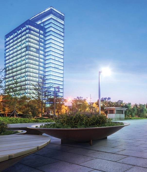 Alt-Text: Modern gestalteter Pflanzbehälter in einem urbanen Park mit einem hohen, gläsernen Gebäude im Hintergrund.