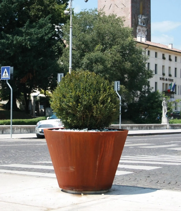 Alt-Text: Großer, konisch geformter Pflanzbehälter aus rostfreier Metalloptik mit einem runden, grünen Strauch in der Mitte, platziert auf einem Straßenabschnitt.