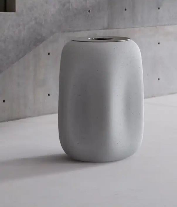Ein moderner, grauer Abfallbehälter aus Beton mit einer geschwungenen Form und einem glänzenden, goldfarbenen Oberteil. Der Hintergrund ist aus schlichtem Beton.