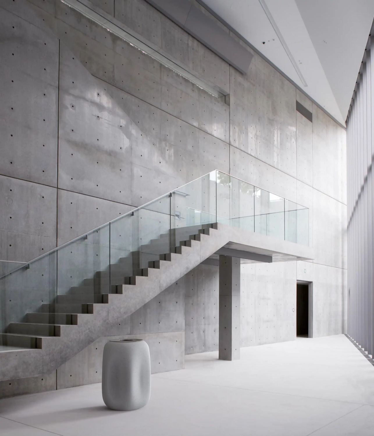 Ein minimalistischer Innenraum mit einer modernen Betontreppe, großen Fenstern und einer glatten, hellen Wand. Ein runder, grauer Abfallbehälter steht am Fuß der Treppe.
