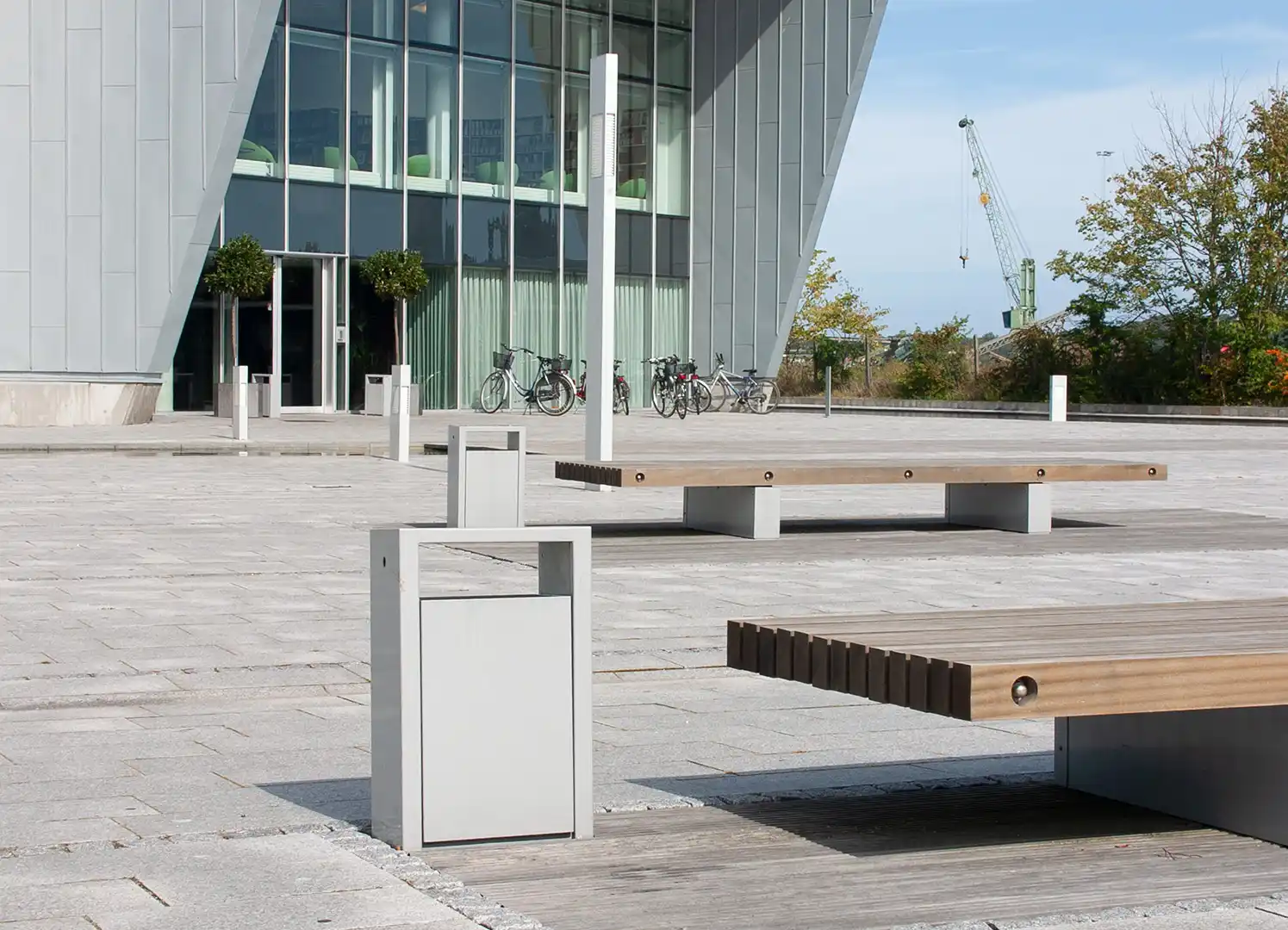 Eine moderne Architektur mit einer Glasfassade im Hintergrund. Im Vordergrund sind Sitzbänke aus Holz und ein Abfalleimer aus Metall zu sehen. Der Platz wirkt ruhig und einladend, umgeben von Bäumen und Fahrrädern.
