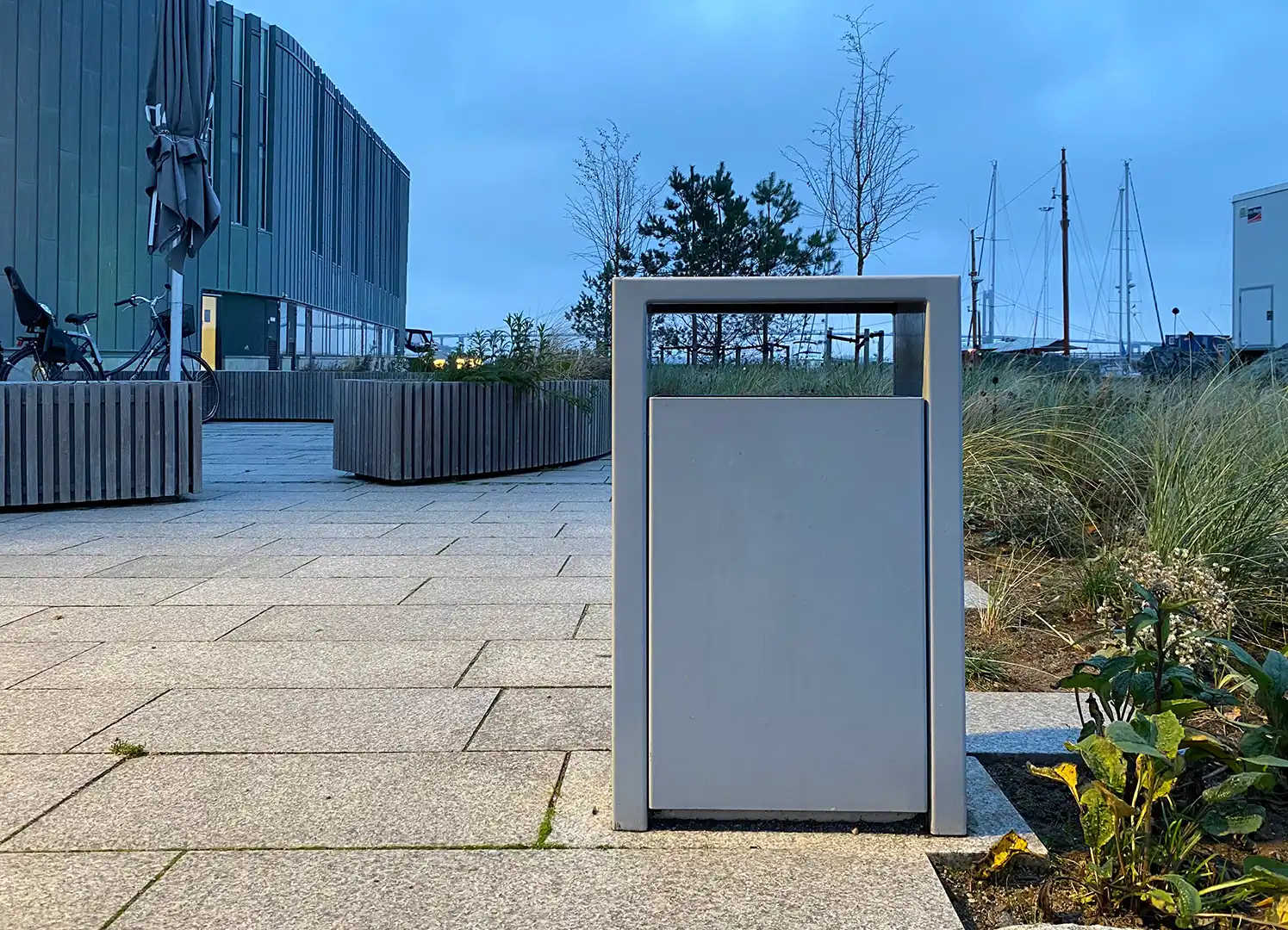 Moderner Mülleimer in einer urbanen Umgebung mit Kieswegen und Pflanzen, im Hintergrund sind Gebäude und Boote zu sehen. Der Himmel ist bewölkt und es ist Dämmerung.