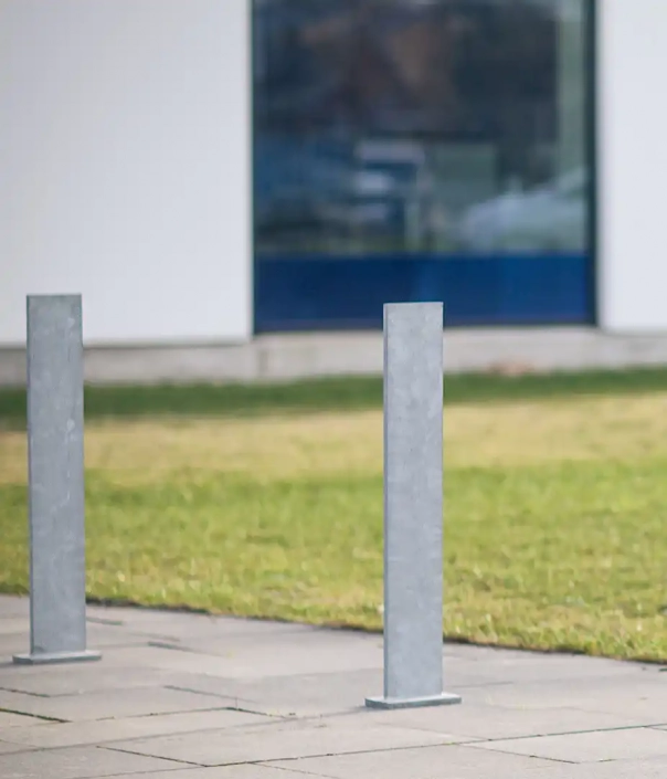 Zwei graue Poller aus Metall stehen auf einer gepflasterten Fläche vor einem modernen Gebäude. Im Hintergrund ist eine grüne Wiese und eine große Fensterfront zu sehen.