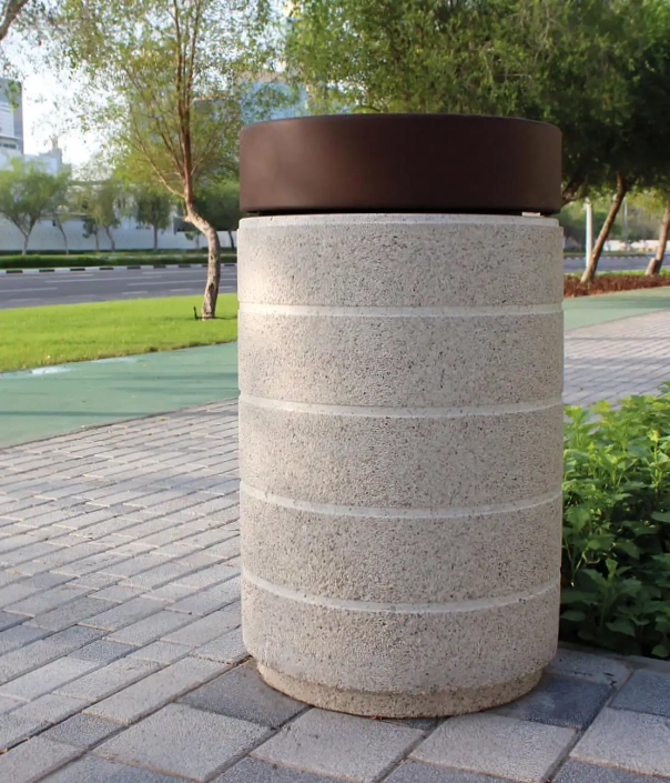 Grauer, zylindrischer Müllbehälter mit einer dunklen, runden Abdeckung, umgeben von gepflegtem Grün und einem Bürgersteig.