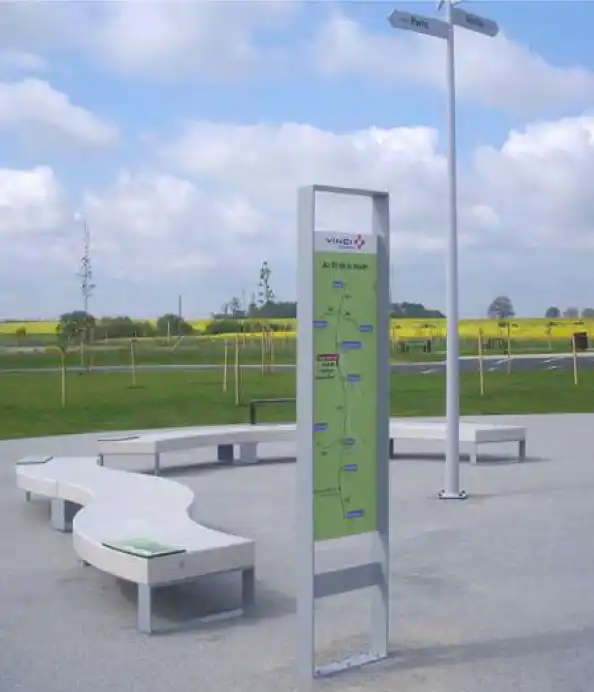 Alt-Text: Übersichtliche Hinweisschilder und eine Informationssäule in einem Park, umgeben von Sitzgelegenheiten und einer grünen Landschaft.