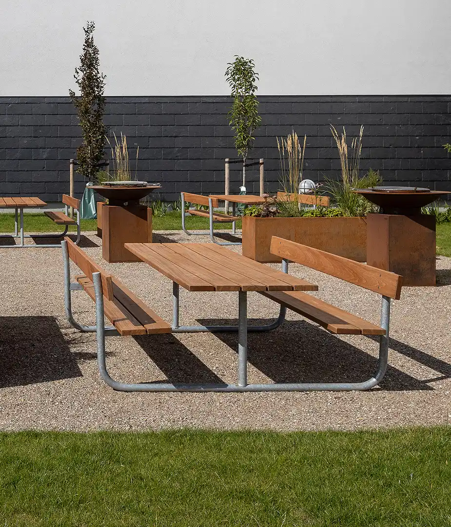 Alt-Text: Sitzgruppe aus Holz und Stahl in einem urbanen Raum mit Grills und Pflanzenbeeten im Hintergrund.