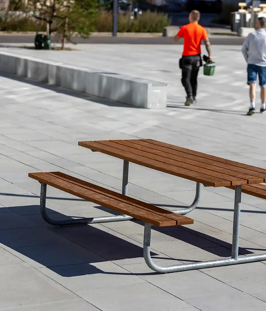 Alt-Text: "Holztisch mit Bänken in urbanem Raum, umgeben von Gehwegen und modernem Design. Im Hintergrund sind zwei Personen zu sehen, die gehen."