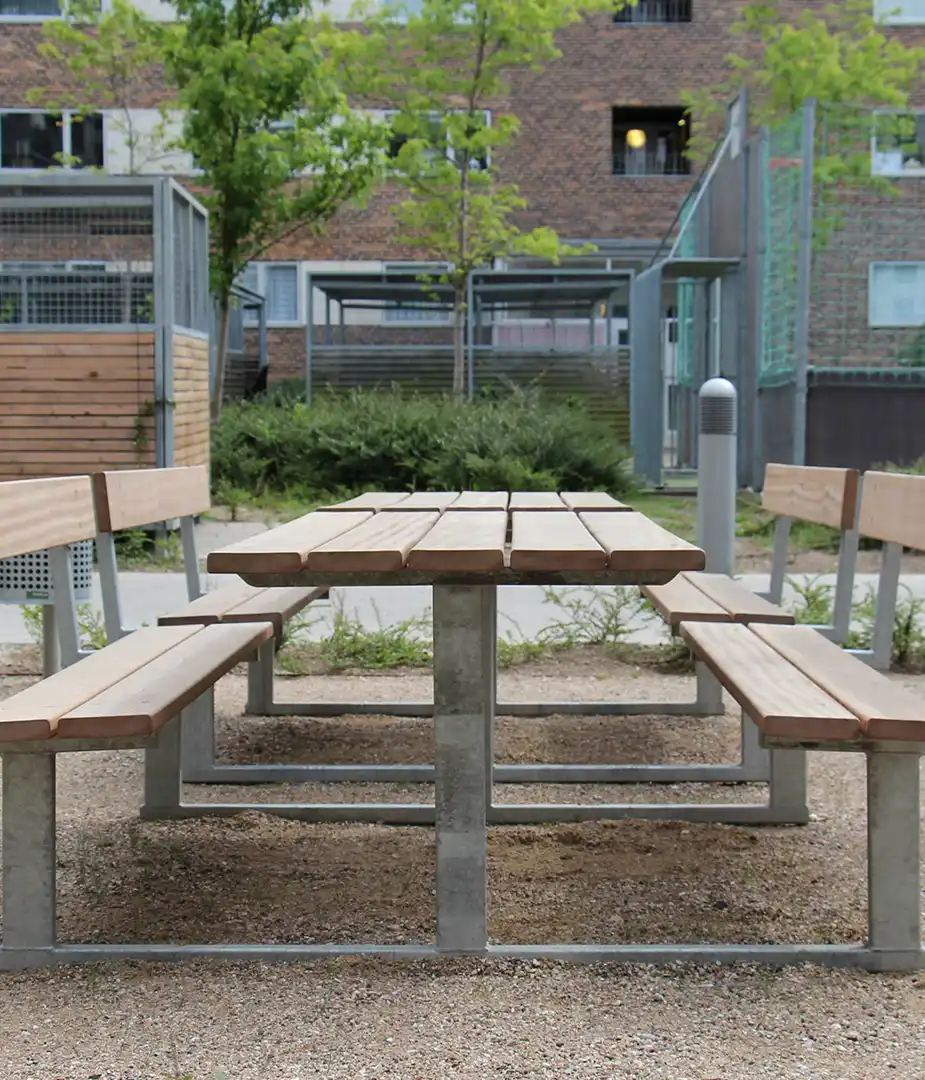 Alt-Text: Sitzgruppe aus Holz mit Tisch und Bänken in einem urbanen Außenbereich, umgeben von Gräsern und Bäumen.
