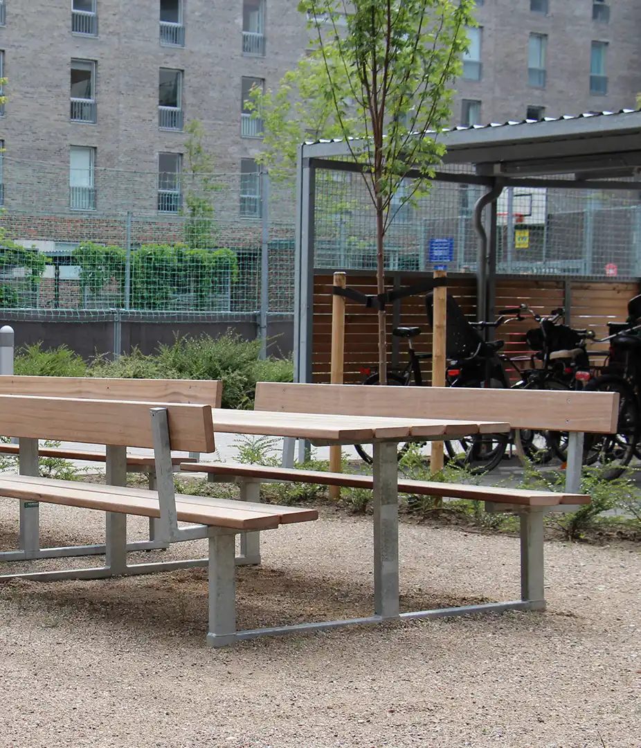 Alt-Text: Sitzgruppe aus Holz und Metall in einem urbanen Aufenthaltsbereich, umgeben von Pflanzen und Fahrradabstellplätzen.