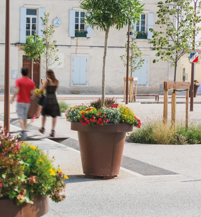 Alt-Text: Große Pflanzbehälter in einer urbanen Umgebung, gefüllt mit bunten Blumen, umgeben von Gehwegen und Passanten.