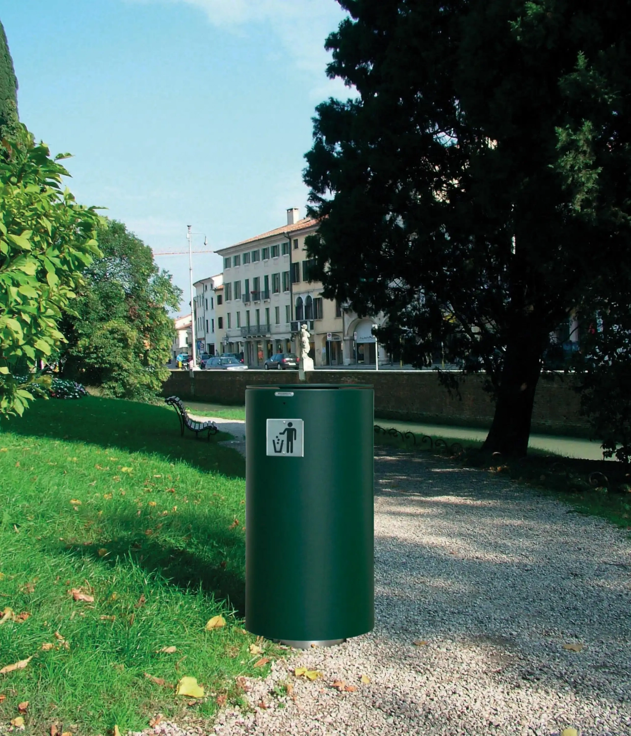 spencer-abfallbehaelter-13 Grüner Abfallbehälter auf einem schmalen Weg, umgeben von Rasen und Bäumen, mit städtischen Gebäuden im Hintergrund.