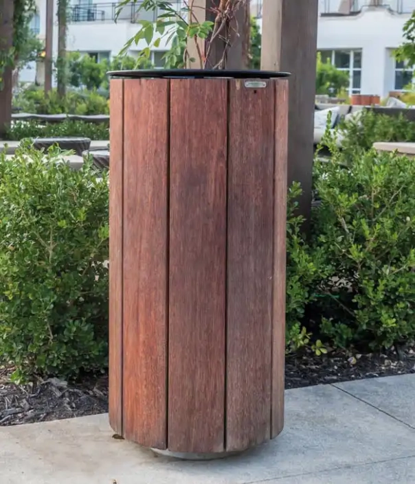 Ein runder Abfallbehälter mit Holzverkleidung vor einem gepflegten Beet.