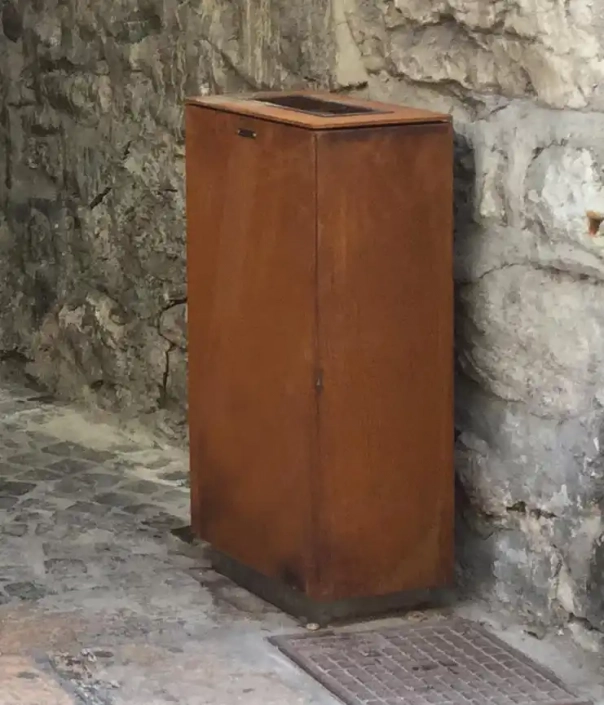 Ein rostfarbener Abfallbehälter vor einer Steinwand. Seine Form ähnelt der eines Kastens und hebt sich durch seine Farbgebung deutlich vom Hintergrund ab.