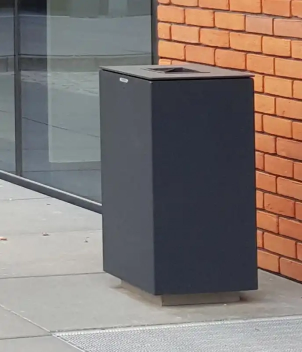 Alt-Text: Eine schwarze Mülltonne steht vor einer modernen, mit roten Ziegelsteinen verkleideten Wand. Der Boden besteht aus grauen Platten.