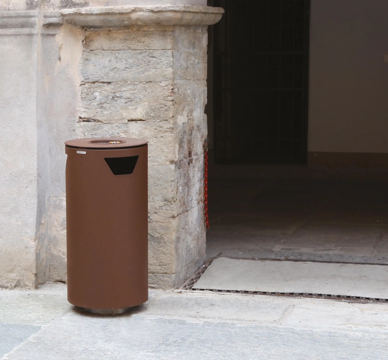 Ein rostfarbener, runder Abfallbehälter vor altertümlichen Mauernweg.