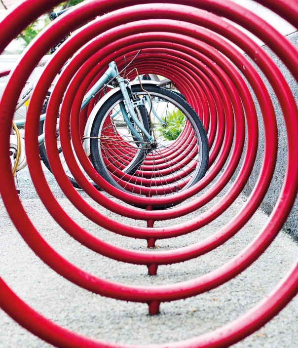 spyra-radverkehr-07 Alt-Text: "Ansicht eines roten Fahrradständers in spiralförmiger Anordnung mit einem Fahrrad im Hintergrund."