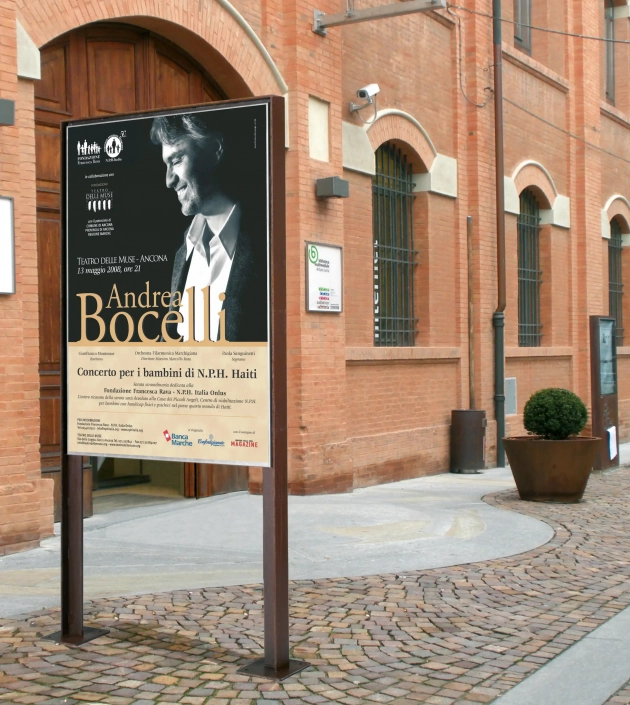 targa-hinweistafel-06 Alt-Text: Ein Hinweisschild für ein Konzert von Andrea Bocelli mit Informationen zum Veranstaltungsort und Datum, aufgestellt vor einem historischen Gebäude mit Backsteinfassade.