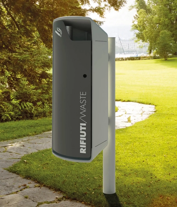 Abfallcontainer in einer Parklandschaft, auf einem Gehweg, umgeben von Bäumen und Gras. Der Container trägt die Aufschrift „RIFIUTI/WASTE“.