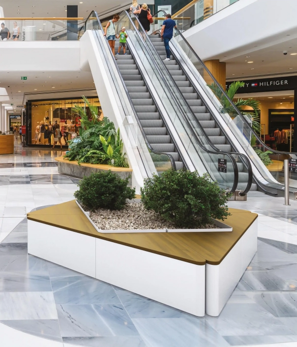 Alt-Text: Moderner Pflanzbehälter aus weißen Materialien mit einer Holzoberfläche, umgeben von grünen Pflanzen, in einem Einkaufszentrum neben einer Treppe.