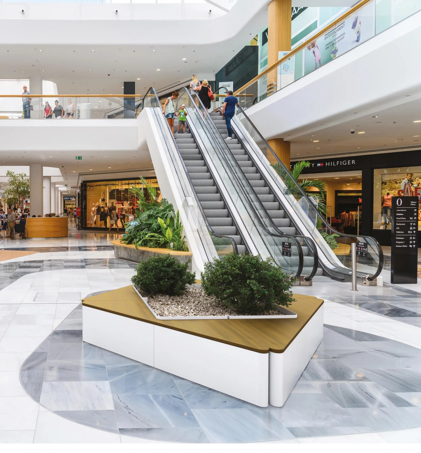 Alt-Text: „Moderne Pflanzbehälter in einem Einkaufszentrum, umgeben von grünen Pflanzen und einer ansteigenden Rolltreppe im Hintergrund."