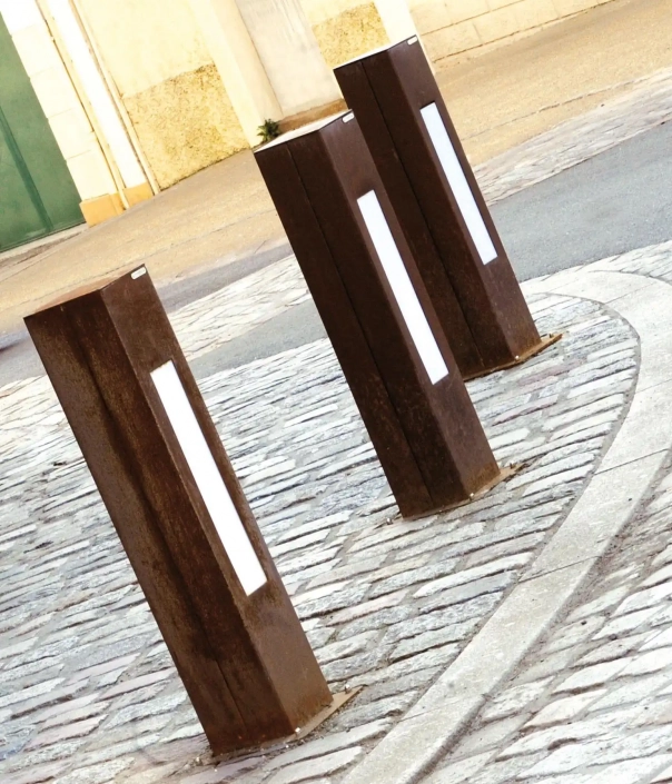 Drei rechteckige Poller aus rostigem Metall, die auf einem gepflasterten Boden stehen, mit weißen reflektierenden Streifen in der Mitte.