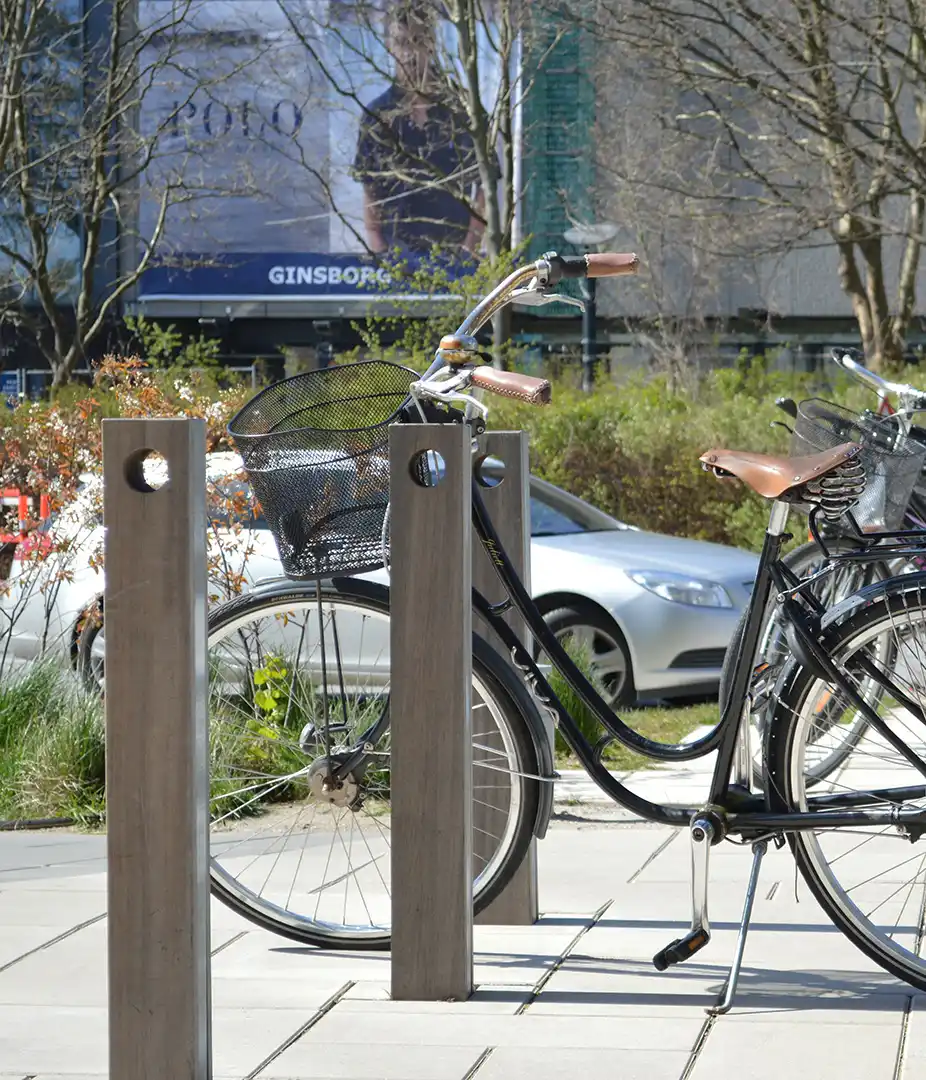 Alt-Text: "Nahaufnahme eines Fahrradständers aus Holz mit einem geparkten schwarzen Fahrrad, ausgestattet mit einem Korb und einem Pumpenanschluss in einem urbanen Umfeld."