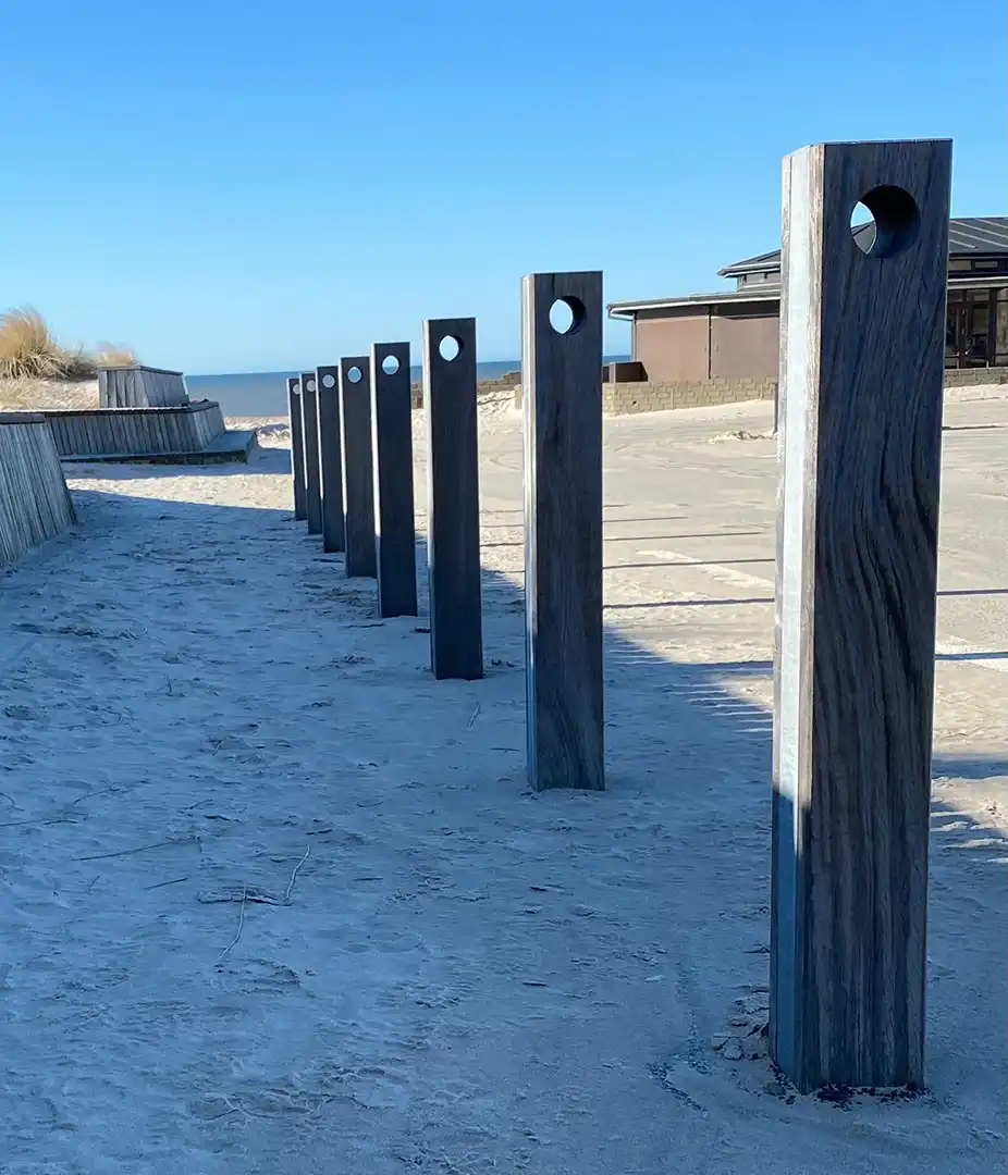 Alt-Text: Eine Reihe von hölzernen Stelen auf einem Sandweg, die als Fahrradständer dienen, mit einem klaren Himmel im Hintergrund.