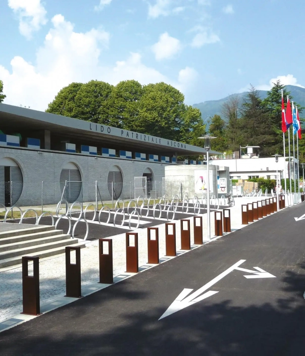 Poller entlang einer Straße vor dem Lido Patriziale in Ascona, umgeben von Bäumen und Flaggen.