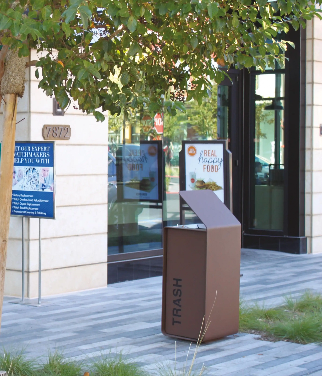 yes-abfallbehaelter-08 Moderner Abfallbehälter mit der Aufschrift "TRASH" vor einem Geschäftsgebäude, umgeben von Pflanzen und einem Baum.