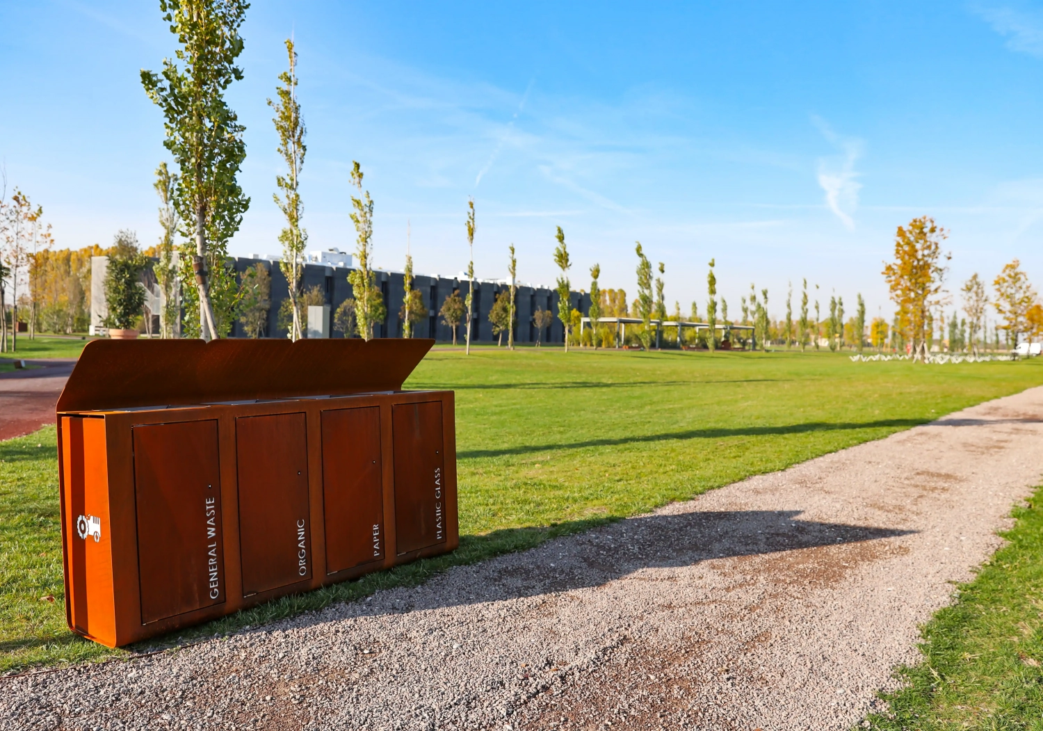 yes-abfallbehaelter-10 Ein Abfallbehälter für organischen Müll steht auf einer Wiese, umgeben von jungen Bäumen. Im Hintergrund ist ein modernes Gebäude zu sehen. Der Himmel ist klar und blau.