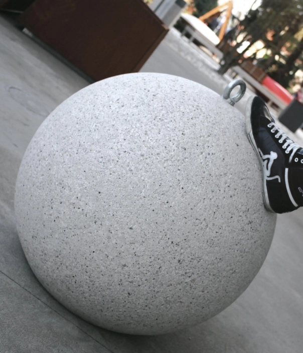 Ein grauer, runder Poller aus Stein, auf dem ein Schuh steht. Hintergrund mit verschwommenen urbanen Elementen.