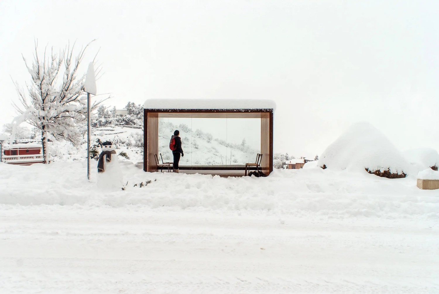Eine moderne Überdachung aus Glas und Holz in einer verschneiten Landschaft, mit einer Person, die darunter steht. Schneeverhangene Bäume und ein verschneiter Weg sind im Hintergrund sichtbar.